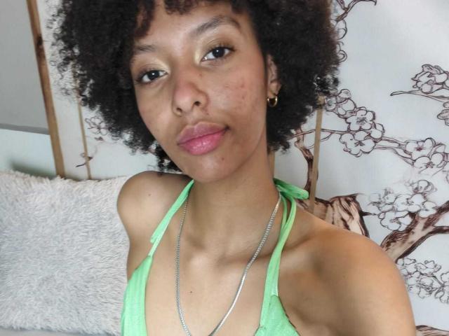 Kiara Kamau, con i capelli neri ricci, la pelle marrone, le labbra rosa, una canottiera verde, piccoli orecchini dorati e una collana d'argento, che mostra le spalle scoperte e la parte superiore del petto..