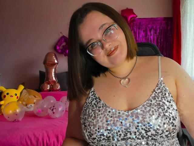 JessicaJoye with shoulder-length brown hair, glasses, and a heart necklace, wearing a silver sequined dress, sitting in front of a pink table with balloons and a Pikachu toy.