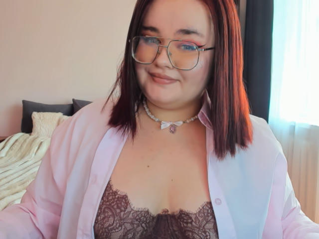 Jessica Joye, avec ses cheveux roux, ses lunettes et son sourire, porte une chemise rose ouverte sur un soutien-gorge en dentelle, un collier avec un pendentif en forme de cœur, et montre ses seins..