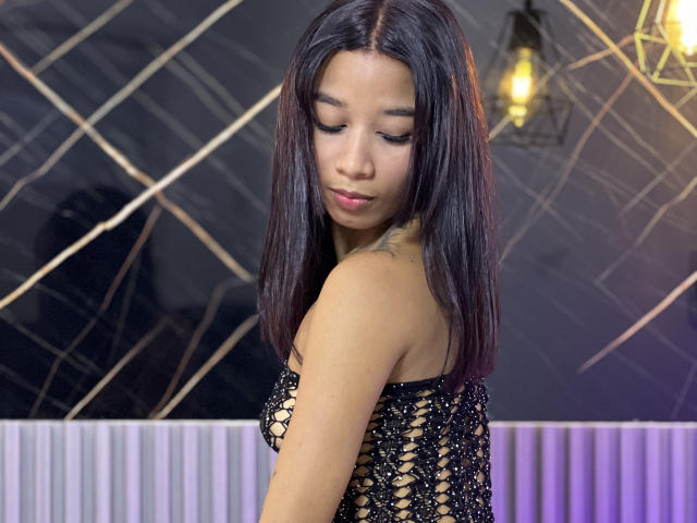 LaurenRosse with long black hair, looking down, wearing a black lace-up dress, exposing her tits, small tattoo on her right shoulder, black diamond-patterned wall, yellow hanging light.