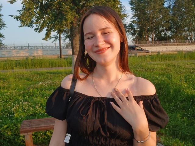 GenevieBidgood with light brown hair, smiling, eyes closed, wearing a black off-shoulder top, touching her chest, hoop earrings, silver necklace, sitting on a wooden bench in green grass.