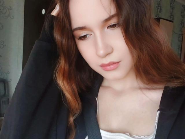 GenevieBidgood with brown wavy hair, light skin, and pink lips, wearing a black hoodie, looking down, has small tits, and a white top with a zipper.