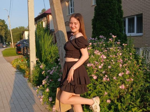 GenevieBidgood, standing with one leg up, has black puffy-sleeve crop top, black ruffle skirt, and white sneakers. She's smiling, with pink flowers and greenery around.