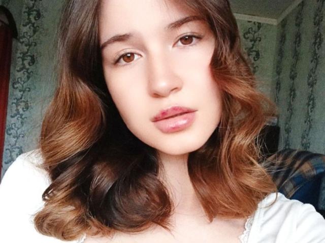 GenevieBidgood has wavy brown hair, brown eyes, and shiny pink lips. She's wearing a white top, and her face is centered in the image.