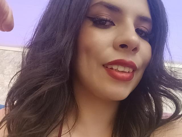 VioletaStone ha i capelli neri e mossi, rossetto rosso e eyeliner con ciglia lunghe. Sorride mostrando i denti, tiene un dito in alto e ha la pelle chiara..