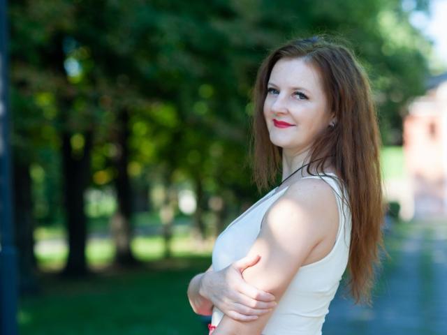 MeganPirs with red hair, white tank top, crossed arms, red lipstick, smiling, green background, standing, looking at camera, cute, tits visible, soft light, outdoor setting.