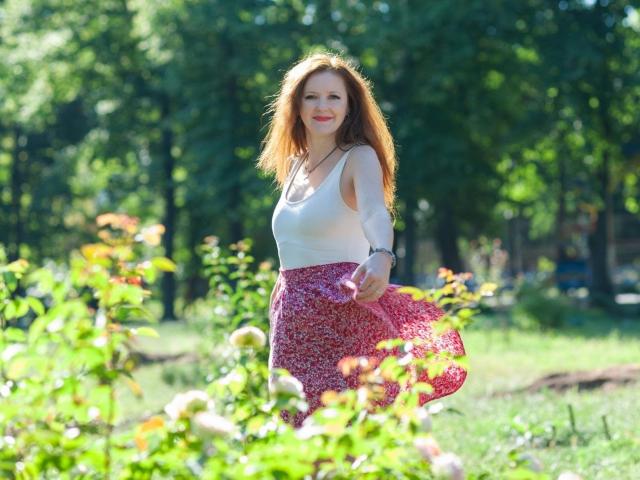 MeganPirs with red hair, white top, and pink floral skirt, standing in a sunlit garden, blowing on her skirt, smiling, with green trees in the background.