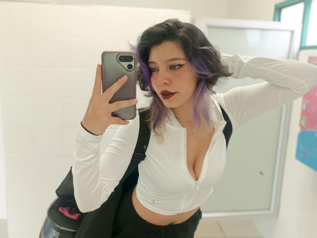 DarckVelvet with purple-dyed hair, dark lipstick, white zip-up top showing her big tits, black vest, black pants, taking a mirror selfie in a bathroom, holding a silver phone.