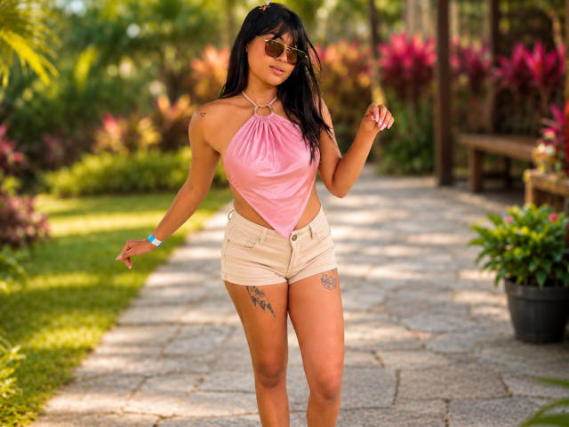 AshlyLee, met een bruine huid, zwart haar, die een roze top met halterneck draagt, beige shorts, tatoeages op haar dijen en een zonnebril, staat op een stenen pad en kijkt naar beneden..