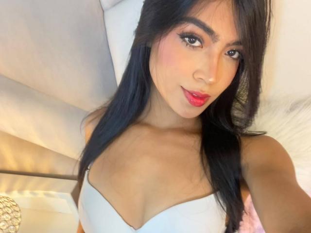CarolineSantos has dark long hair, red lipstick, and is wearing a white strapless top showing her small tits. She's lying on a white couch with a gold pillow.