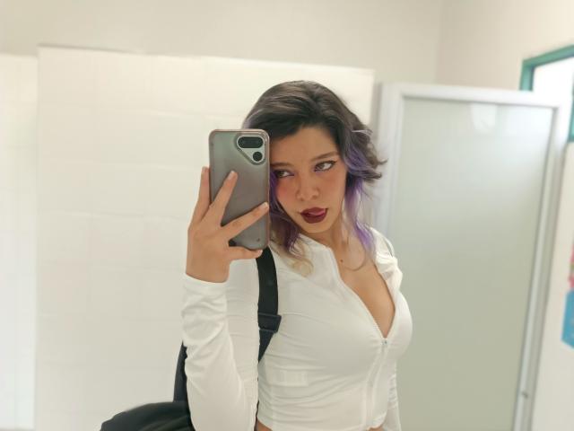 DarckVelvet with purple-tipped hair, wearing a white, low-cut shirt, showing her tits, holding a phone, taking a mirror selfie, with a black backpack.