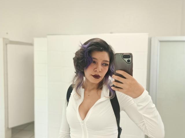 DarckVelvet with purple-dyed hair, dark lipstick, white zip-up top, and black backpack, taking a mirror selfie with a smartphone in a bright, white-walled room.