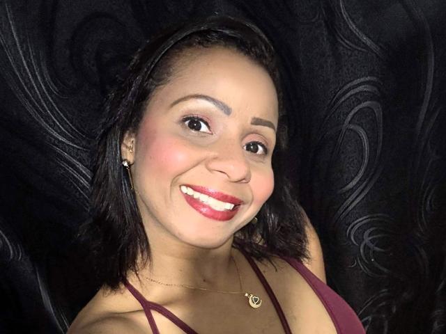 AlondraMonique, con la pelle marrone scura, i capelli neri lunghi fino alle spalle, il rossetto rosso, che sorride, indossa una maglia color borgogna, una collana dorata e orecchini, su uno sfondo nero con motivi..