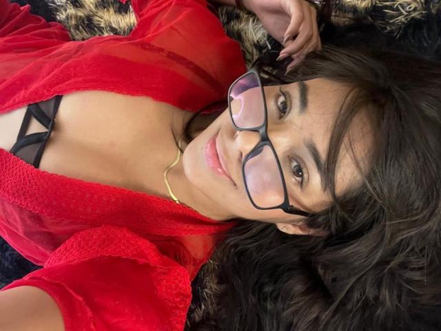 Sofia Cardenas, allongée sur un lit, portant une robe rouge transparente, un soutien-gorge noir, des lunettes et un collier en or, souriant à la caméra, avec ses cheveux bruns ondulés et ses yeux marrons..