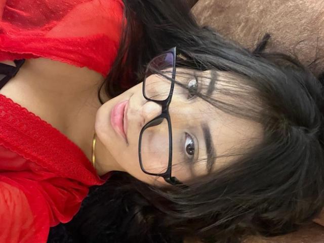 SofiaCardenas lying on a brown surface, wearing black glasses and a red lace top, showing her boobs, with black hair and a subtle smile.