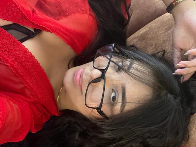 SofiaCardenas with black glasses, red lace top, and black bra, lying down, showing her tits, long black hair, and a gold necklace, smiling at the camera.