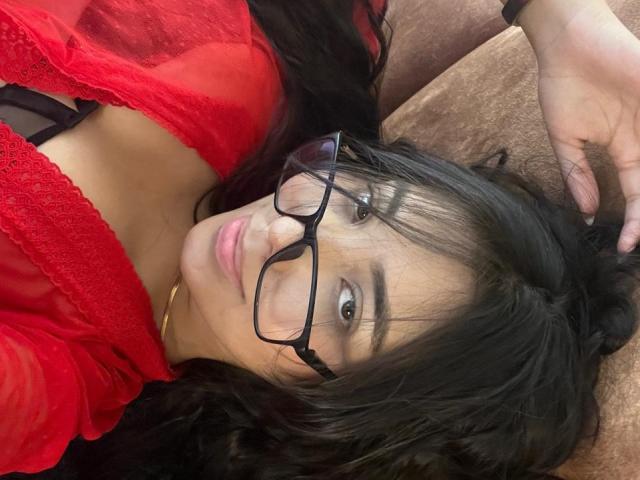 SofiaCardenas with black glasses, red lace top, and black bra, lying on brown couch, hand in hair, looking at camera, soft smile, wavy black hair, light brown skin.