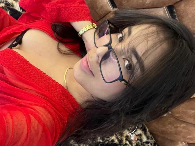 Sofia Cardenas, avec des lunettes, en robe rouge en dentelle, montrant ses seins, allongée sur du cuir marron, cheveux noirs, montre en or, sourire discret, lumière douce..