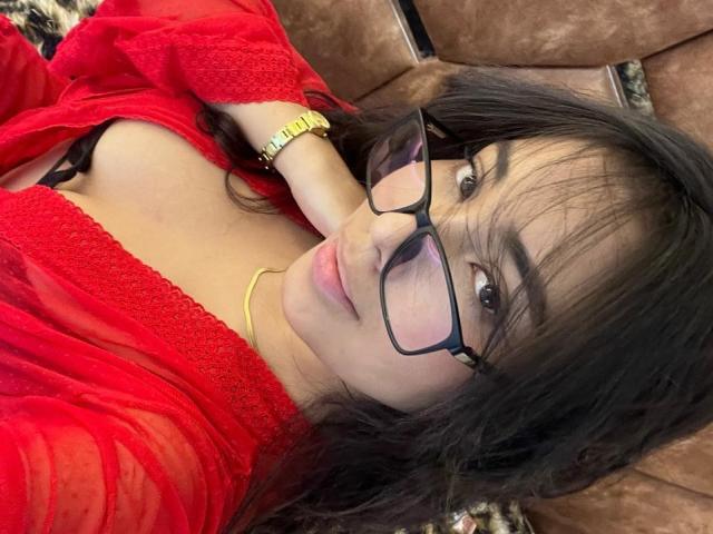 Sofia Cardenas, avec ses cheveux foncés, ses lunettes, sa robe en dentelle rouge, son bracelet jaune et ses lèvres roses, allongée sur un canapé marron, montrant ses seins, regardant la caméra et souriant légèrement..