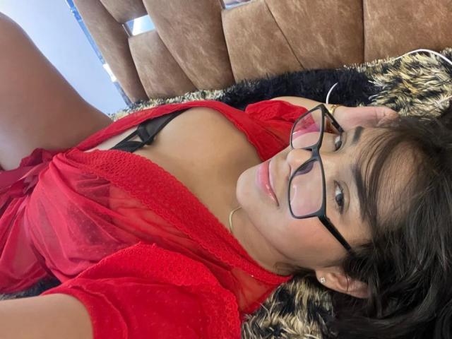 SofiaCardenas with glasses, lying on a bed, red sheer top, black lace bra, exposing her small tits, smiling, brown hair, black and brown fur blanket.
