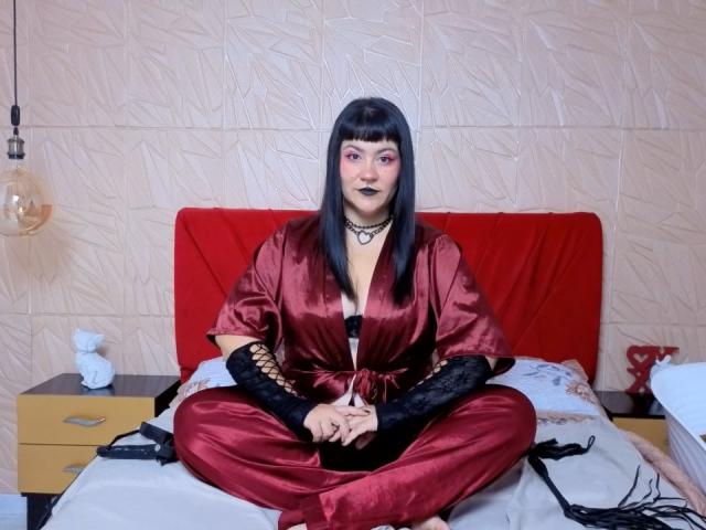 NaniSky mit schwarzen Pony, trägt einen roten Seidenbademantel, schwarze Spitzenhandschuhe und rote Hosen, sitzt mit angelegten Beinen auf einem Bett, die Hände liegen im Schoß, trägt dunkelroten Lippenstift und eine schwarze Halskette..