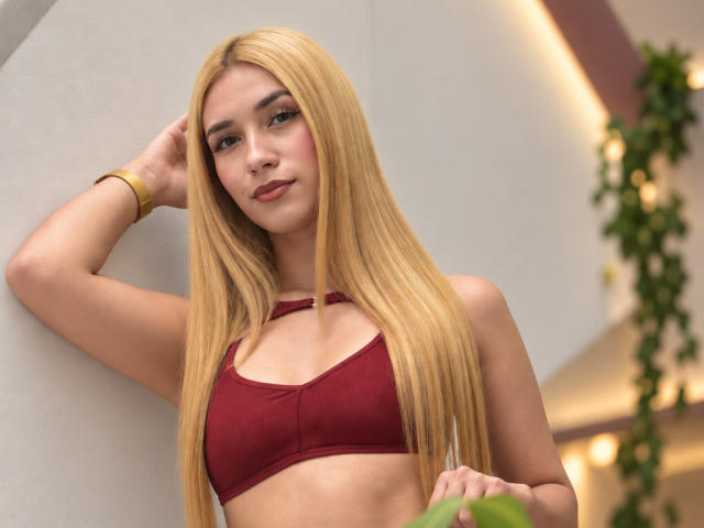 TanniaMoon, con cabello rubio largo, vestida con un top deportivo rojo, tiene senos pequeños, una cara linda y una pulsera dorada, y está de pie con una mano en la cabeza..