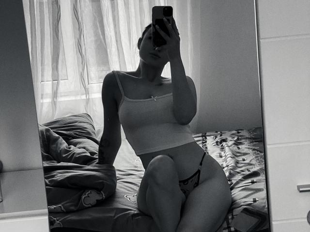 Olieri with small tits, wearing a white crop top and black thong, sits on a bed, taking a mirror selfie, holding a phone, in a grayscale photo.
