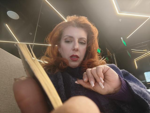 Khharha with red hair, wearing a blue sweater, holding a book, looking down, her hand with long nails near her face, her red lips slightly parted, in a low angle shot.