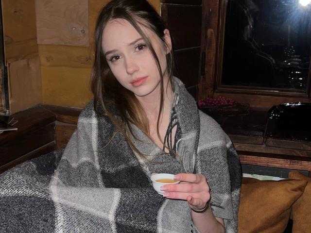 RikiFlowers with long brown hair, wearing a gray and black plaid blanket, holding a small white cup, sitting in a dimly lit room, looking slightly bored.