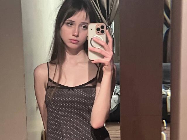 RikiFlowers, with long brown hair, small tits, wearing a black polka dot dress, taking a mirror selfie with a gold iPhone, looking serious, in a dimly lit room.