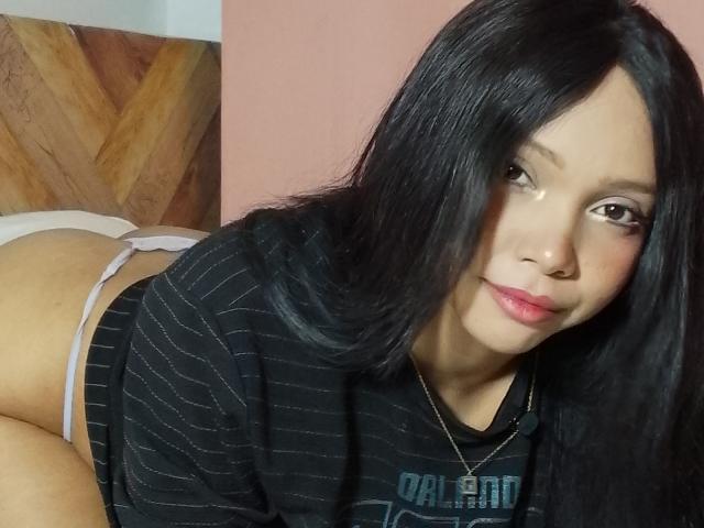 NashaLoule with long black hair, wearing a black striped shirt, white panties, and a necklace, lying down, showing her ass, looking at the camera with a slight smile.