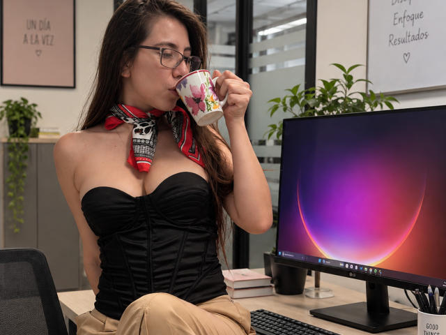 FreyaraVenwo with glasses, sipping from a floral mug, wearing a black strapless corset, red scarf, and tan pants, sitting at a desk with a colorful computer screen.