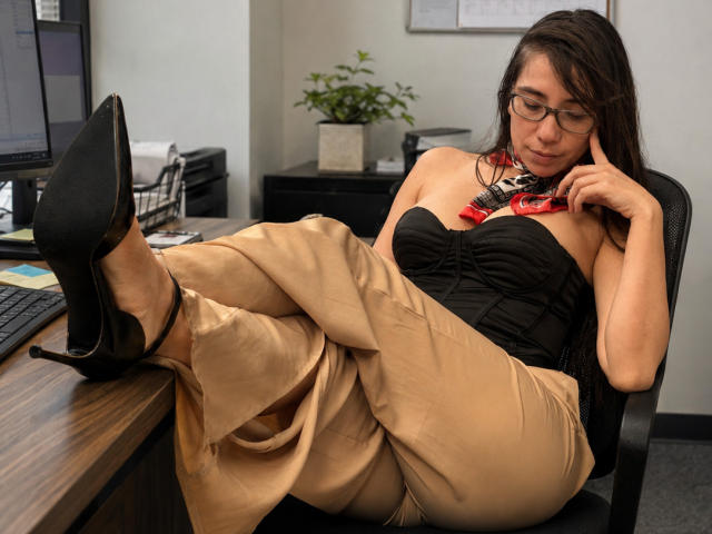 FreyaraVenwo, with glasses and dark hair, lounges in an office chair, legs up, wearing a black strapless top, beige pants, and black heels, resting her chin on her hand.