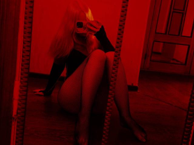 Red-lit photo of TiffaniMacandog sitting on wooden floor, wearing fishnet stockings and black top, legs crossed, blonde hair, mask covering lower face, coiled mirror frame in background.
