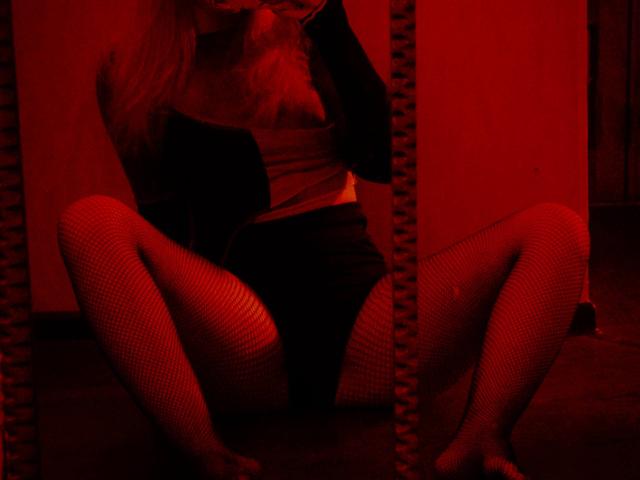TiffaniMacandog sits with legs spread, wearing fishnet stockings and black underwear, her arms covering her tits, bathed in red light. Her ass is prominent in the shot.