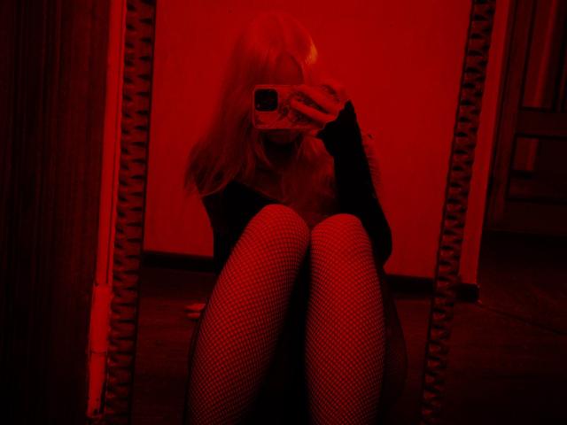 Red-lit mirror selfie of TiffaniMacandog with blonde hair, fishnet stockings, and black long-sleeve top, holding phone covering face, sitting with knees up, showing legs.