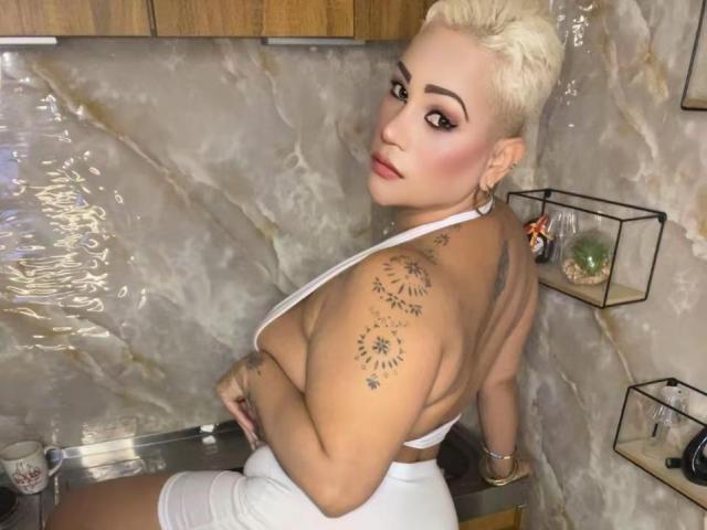 Gianella Cox, com cabelo loiro curto, blusa de alça branca e shorts brancos, exibindo suas tatuagens nas costas e no braço, sentada em uma cozinha, olhando por cima do ombro..