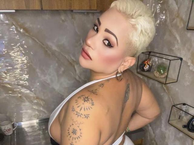 Gianella Cox, avec ses courts cheveux blonds, regarde en arrière, exhibant son dos tatoué et ses grandes seins dans un haut blanc à encolure dégagée. Elle porte des boucles d'oreilles cercles et a un regard séduisant..
