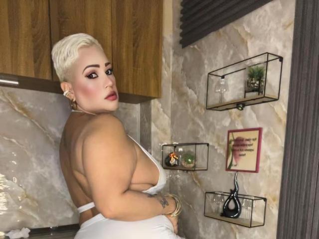 GianellaCox with platinum blonde pixie cut, white backless dress showing big tits and round ass, looking over shoulder, standing in modern room, wooden cabinets, marble walls.