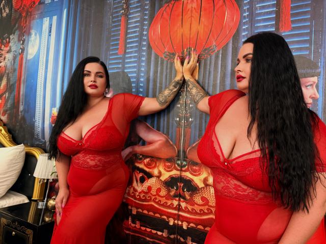 Two GeorgiaLerox women with large tits, black hair, red lace lingerie, and tattoos, standing in front of a red lantern, touching hands, in a richly decorated room.