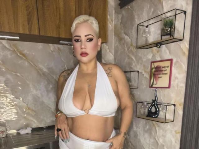 GianellaCox with platinum blonde hair, wearing a white halter top that exposes her big tits and a tattoo on her right shoulder, standing in a kitchen.