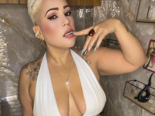 GianellaCox, avec ses cheveux blonds courts, ses seins volumineux, un haut à encolure américaine blanc, des ongles bleu-vert, un tatouage sur l'épaule, touchant sa langue avec un doigt, portant des boucles d'oreilles cerceaux et une bague rouge..