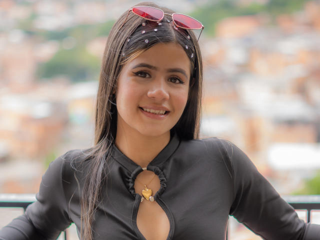 SheylaSpooner with long brown hair, wearing a black top with a keyhole, pink sunglasses on head, smiling, sitting outdoors, background blurred. Boobs visible through top's cutout.