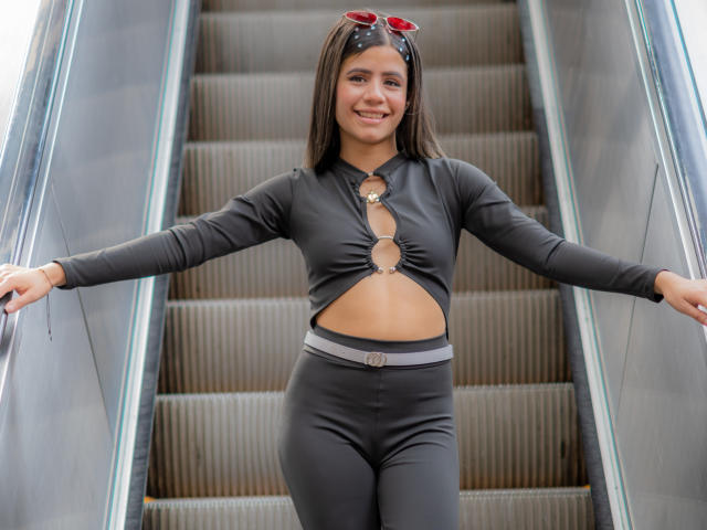 SheylaSpooner with dark brown hair, smiling, wearing a black cut-out long-sleeve top and matching black pants, standing on an escalator, with red sunglasses on her head.