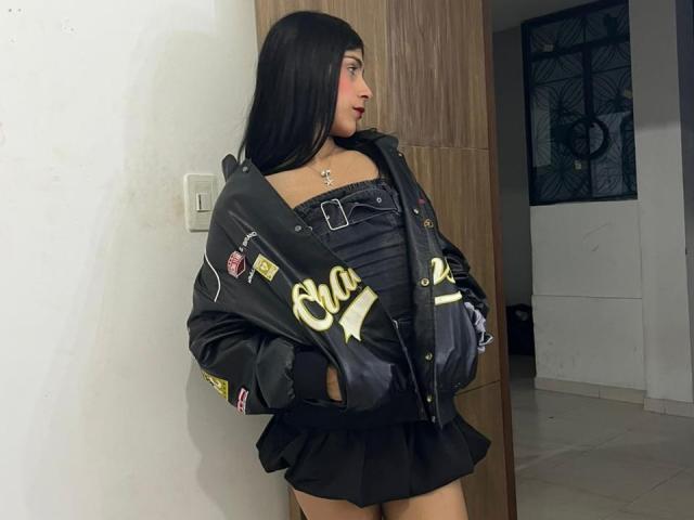 ValeryTranss with long black hair, wearing a black leather jacket, off-shoulder top, and black skirt, standing in a room, hands in pockets, looking to the side.