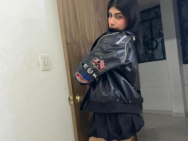 ValeryTranss with long dark hair, wearing a black leather jacket with patches and a black pleated skirt, standing in a doorway, looking back with a slight smile.