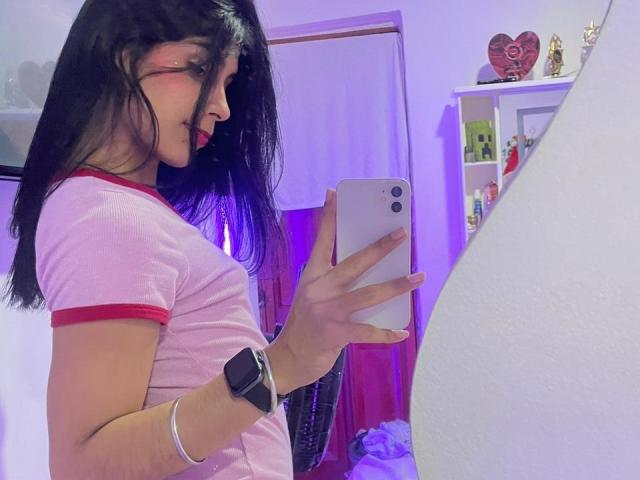 ValeryTranss with long black hair, red lipstick, pink shirt with red trim, holding a white phone, wearing a smartwatch, taking a mirror selfie in a purple-lit room.
