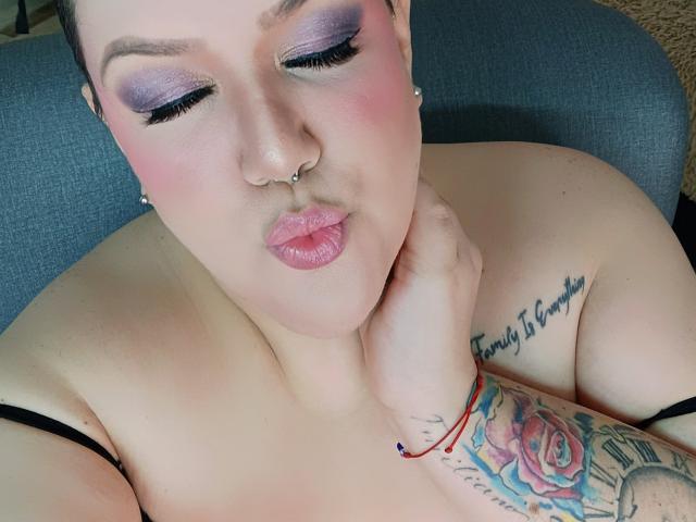 GiiannaMorrys with purple eye makeup, pink blush, nose ring, pouty lips, tattooed arm, rose tattoo, red bracelet, hand on neck, bare shoulders, short hair, blue chair.