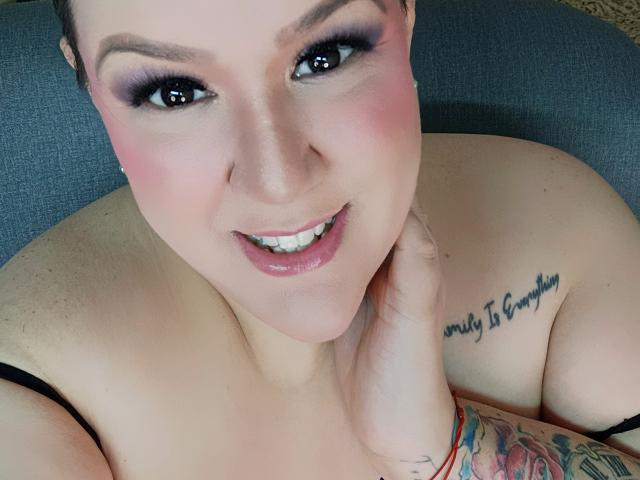 GiiannaMorrys with pink eyeshadow, glossy lips, and a tattoo reading "Never 2 Gonna" on her shoulder, touching her chin. Her tits are visible, and she has a colorful arm tattoo.