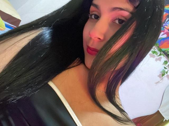 ValeryTranss with long black hair, pink highlights, red lipstick, and a black top showing off her tits. She's looking at the camera with a slight smile.